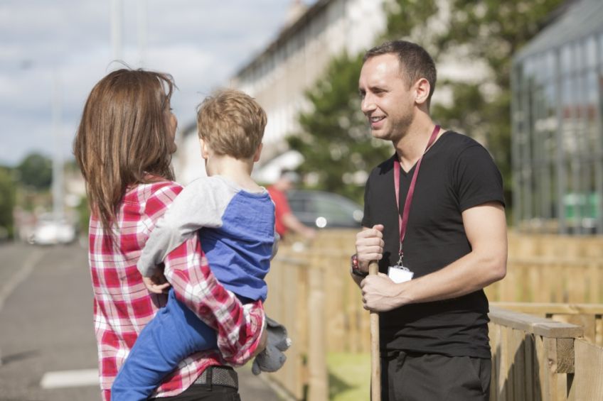 A male housing officer talking to a female customer holding a small child on the pavement next to houses